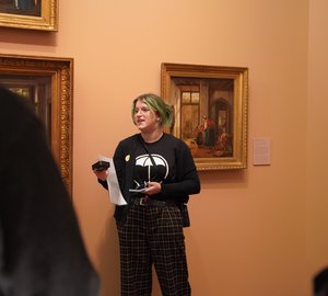 Take pARTner Flame geeft een rondleiding, Flame staat met een papier in haar hand en verteld enthousiast aan toeschouwers over een schilderij.