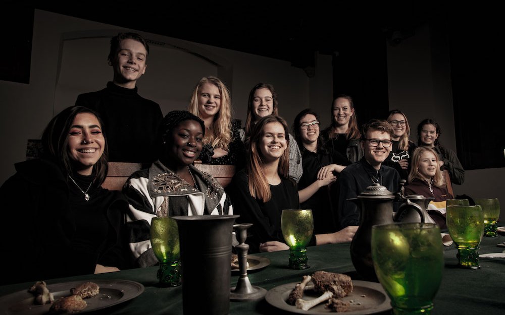 De lachende jonge mensen zitten aan een gedekte tafel. De sfeer is een beetje donker, net als in vroegere tijden. Deelnemers van TakepART 2018-2019.